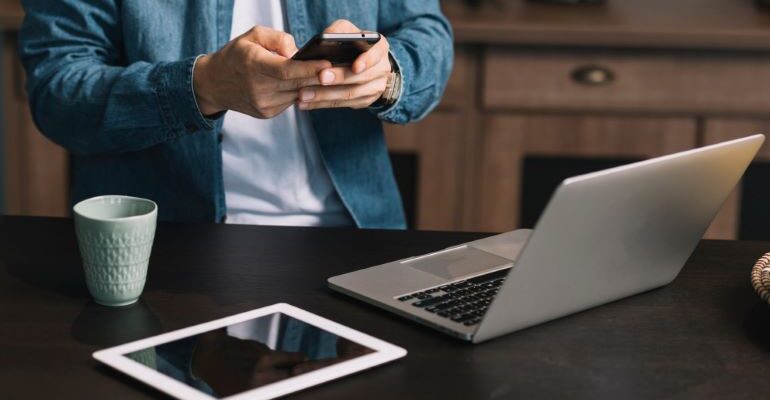 Person browsing on his phone with a laptop and tablet in front of him.