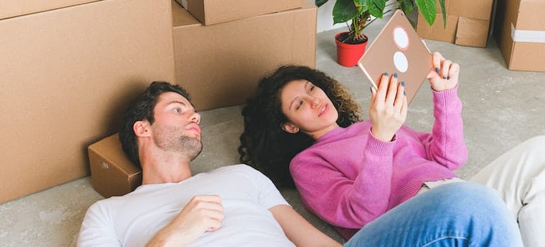 A man and a woman lying on the floor and looking at a photo, surrounded by moving boxes.