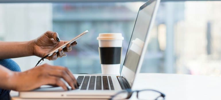 A person typing on their laptop while looking on their phone and researching web design practices for moving companies. 