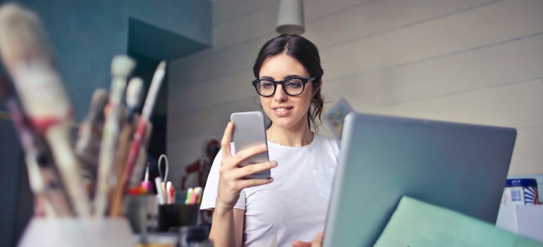 A woman using her smartphone while working on a laptop, illustrating the use of social media in promoting self-storage services.