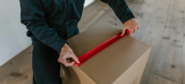 A person sealing a moving box with red tape, illustrating how market shifts and your moving business require adaptability and efficient packing services.