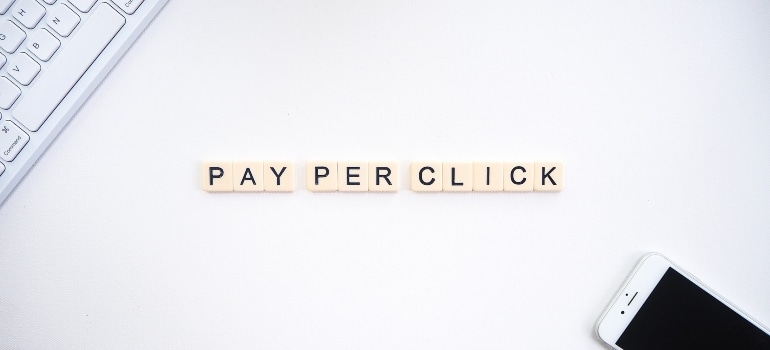 Scrabble tiles spelling out 'PAY PER CLICK' on a white background, with a keyboard and smartphone nearby.