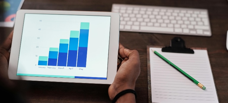 Hands holding a tablet displaying a bar graph, with a notebook and keyboard in the background.